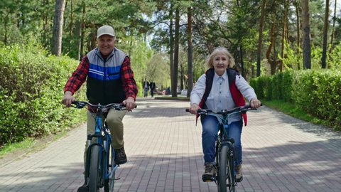 Tracking of cheerful elderly woman and senior man learning to ride bicycles in green park on summer day