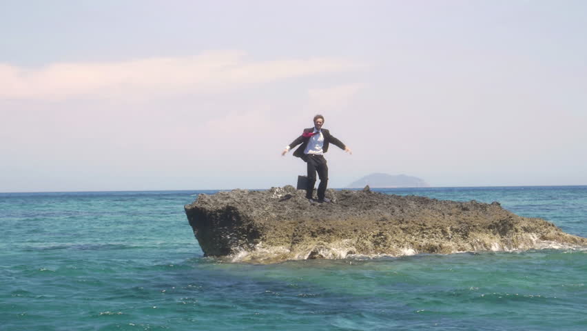 Man in suit whit a briefcase stranded on a deserted rock in the middle of the ocean waving his hands and moving around calling for help