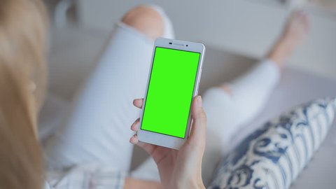 Young Woman in white jeans laying on couch holds SmartPhone with pre-keyed green screen. Perfect for screen compositing. Made from 14bit RAW. 10bit ProRes 444