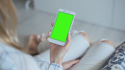 Young Woman in white jeans laying on couch holds SmartPhone with pre-keyed green screen. Perfect for screen compositing. Made from 14bit RAW. 10bit ProRes 444