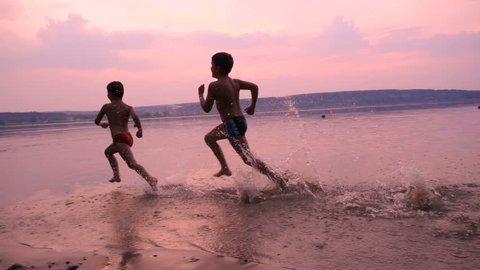 Silhouette of two boys running together on river's beach against sunset, slow motion 스톡 비디오