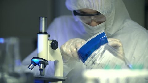 Forensic crime scientist places evidence sample on microscope to analyse clues in laboratory. White Caucasian Male wearing protective clothing for non-contamination. Police murder investigation.