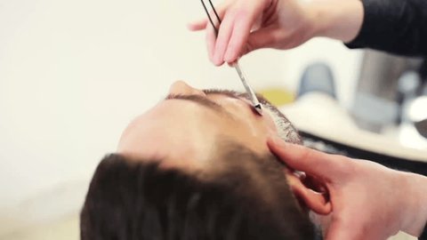 beauty, grooming, hairstyle and people concept - man and hairstylist with straight razor shaving beard at barber shop