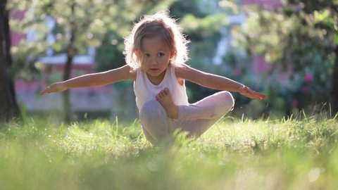 Little cute baby girl doing yoga exercise on the grass at beautiful sun light in green summer park.