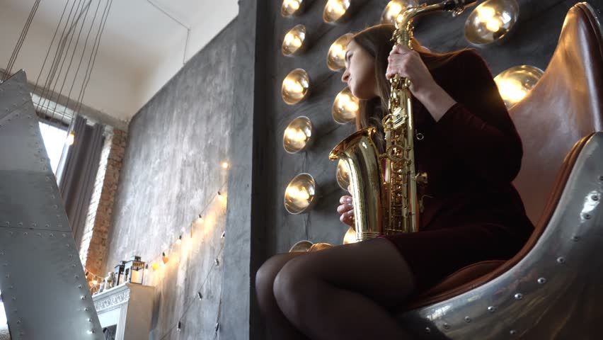 Girl in dress with saxophone sitting on swivel chair in retro room on a background of wall with powerful lamps. Dolly shot