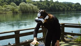 Young Woman Wearing VR Virtual Reality Headset Device and Moving Her Arms in an Outdoor Footpath by the River