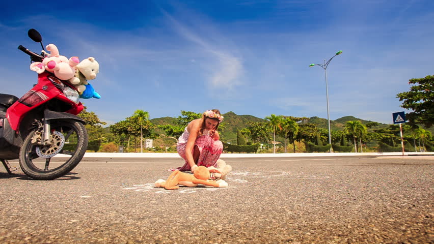 young nice lady in long pink dress and garland squats by red motorcycle and draws on street asphalt. time lapse