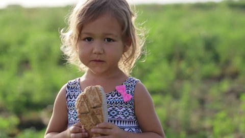 Portrait of cute little girl eating homebaked bread on the field of organic eco farm. Baby holding loaf of bread in hands and funny biting it.