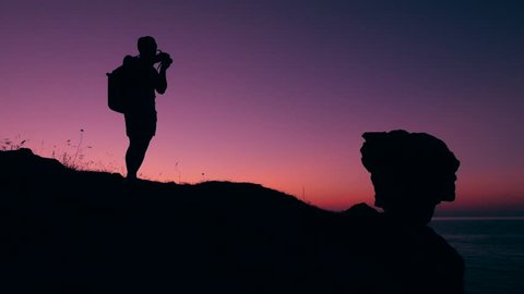 Photographer Taking Photos With Professional Camera on the Mountain Peak. Hiker Tourist With Backpack Silhouette and Amazing Pink Sunset on Background. HD Slowmotion Footage.