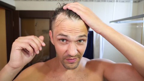 Young attractive man inspects a receding hairline in the bathroom. Baldness. 4k, slow motion
