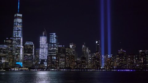 Two beams of light rise up into the New York City skyline at night, as aircraft fly by and boats travel across the Hudson River. 