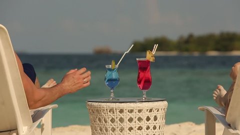 Young family couple sunbathing and drinking colored cocktail at tropical beach. Lovers on honeymoon have exotic vacation lying in the sun on deck chairs near ocean