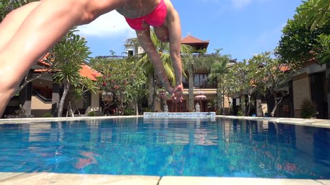 SLOW MOTION CLOSE UP: Young woman on summer vacation jumping head first into blue pool water for refreshment on hot sunny day. Girl in pink bikini diving into clear pool water in empty luxury resort