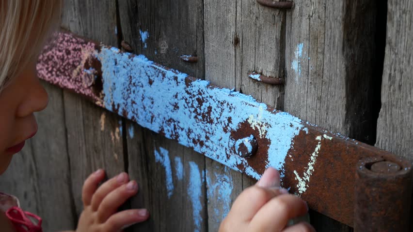A child paints with chalk on an old wooden door. The girl paints a part of the wall with colored crayons.