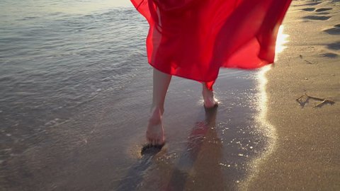 Young barefoot girl in long red dress running dancing along the surf line. Happy girl with long hair having fun on sandy beach during summer holidays. Female bare feet and fluttering dress at sunset.