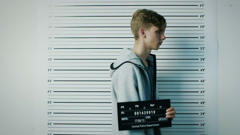 In a Police Station Arrested Teenage Delinquent Steps in for Side, Front View Mugshot. He is Heavily Bruised. Height Chart in the Background. Shot on RED EPIC-W 8K Helium Cinema Camera.