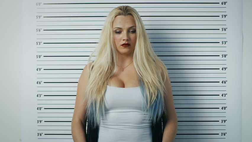 In a Police Station Arrested Woman Posing for Side, Front-View Mugshot. She Chews Gum, Wears Saucy Clothes, Has Smudged Heavy Makeup, Her Hair Is Disheveled and She Holds Placard.Height Chart on Wall.