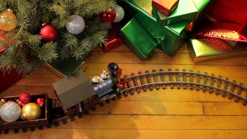 Toy electric train filled with christmas ornaments passes along wood floor on