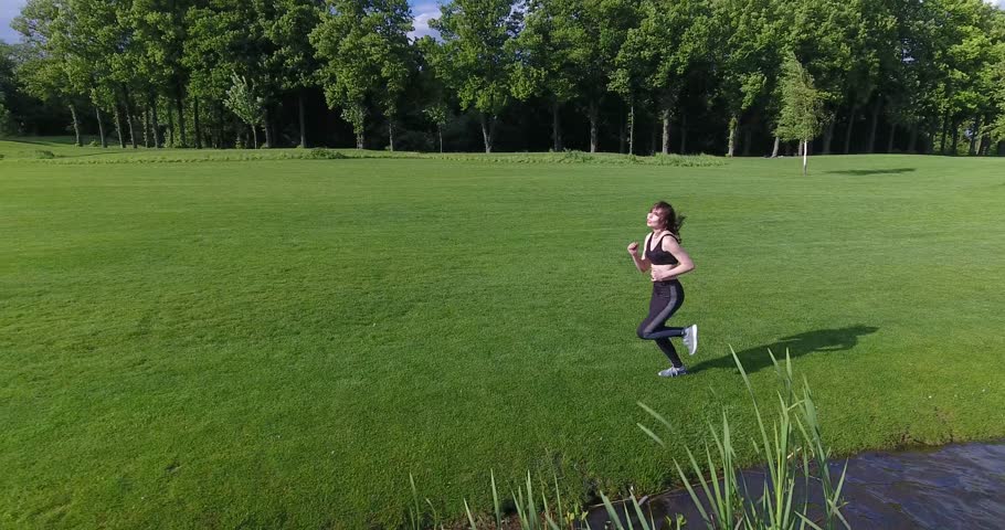The horizontal view of the athletic woman jogging along the meadow near the river. The moving back view.