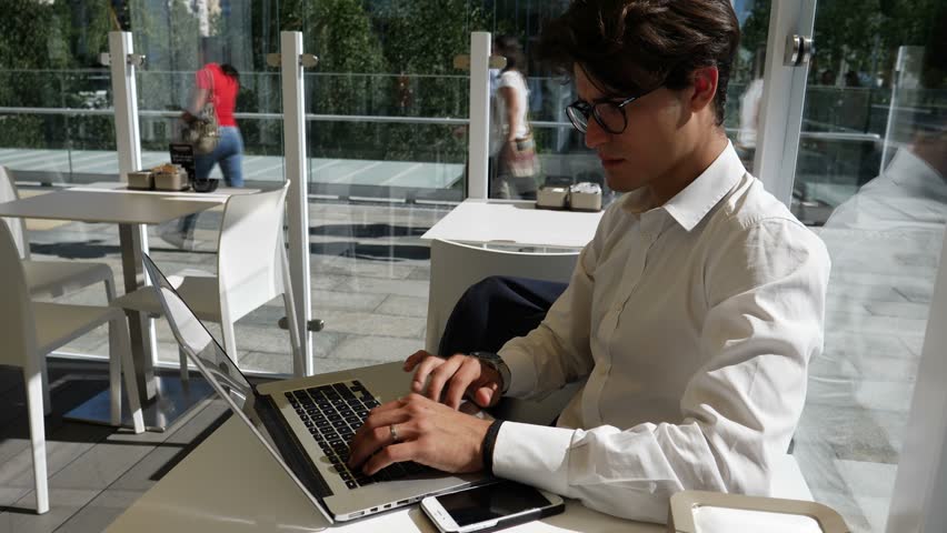 Handsome trendy young business man wearing white shirt working on laptop computer at bar table, outdoor in city setting