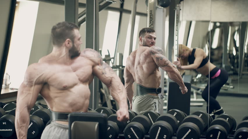 In the gym, a man looks in the mirror and makes swings with muscular arms. A powerful athlete breeds tattooed limbs to the sides. The athletic belt is worn while performing strength exercises with
