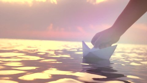 Kid putting a paper boat into water over beautiful sunset. Little boy's hand puts paper ship on sea surface. Origami ship Sailing. Dreams, future, childhood, freedom or hope concept. Slow motion 4K