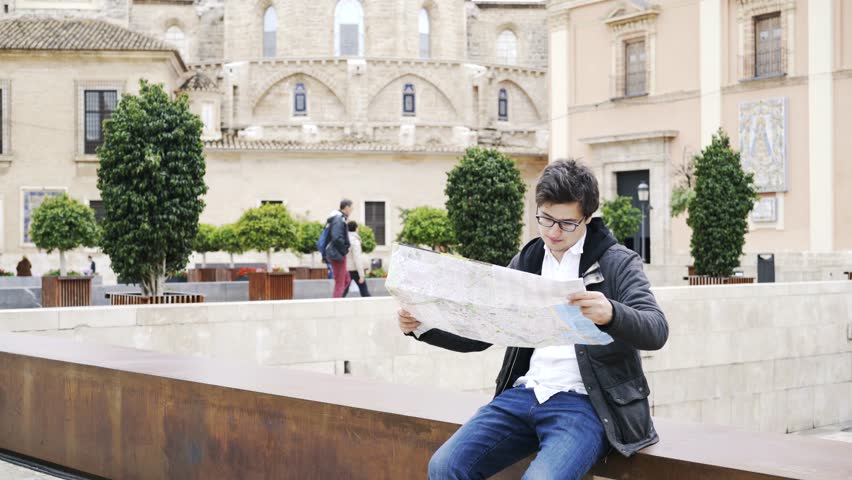 Portrait of a young man in glasses looking at a map he is holding. Valencia, Spain. Beautiful architecture. Locked down real time medium shot