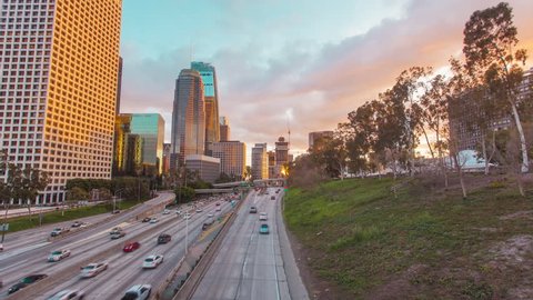 Timelapse in motion (hyperlapse) shot at sunset facing downtown Los Angeles at twilight changing from day to night with beautiful gold and blue skies above and busy street traffic next to trees below.