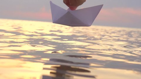 Kid putting a paper boat into water over beautiful sunset. Little boy's hand puts paper ship on sea surface. Slow motion 4K