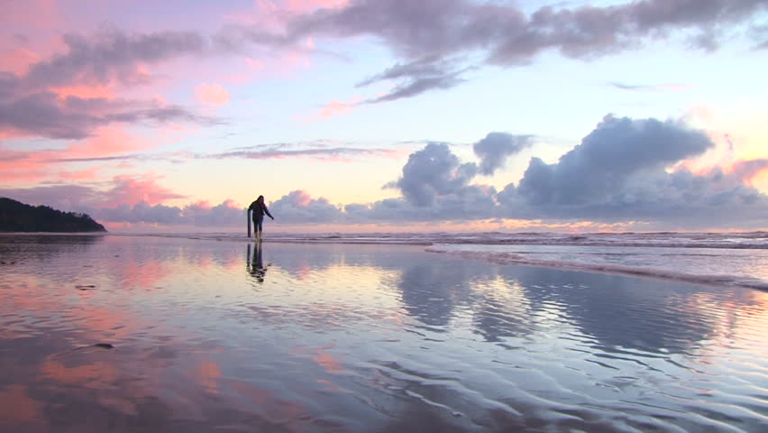 Woman at Pacific Ocean runs from tide coming in on sandy beach at sunset.