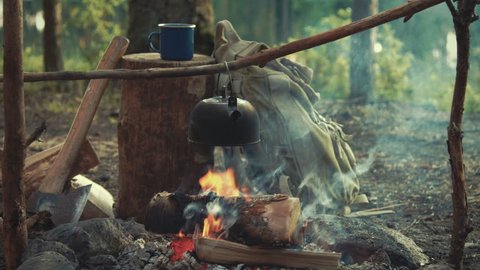 Idyllic camping scene with blue enamel mug, backpack, an axe and tea pot boiling over open fire medium shot