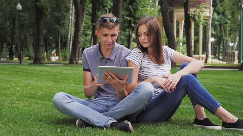 A girl and a guy use a digital tablet. Couple in park sitting on grass watching in digital tablet, touching screen