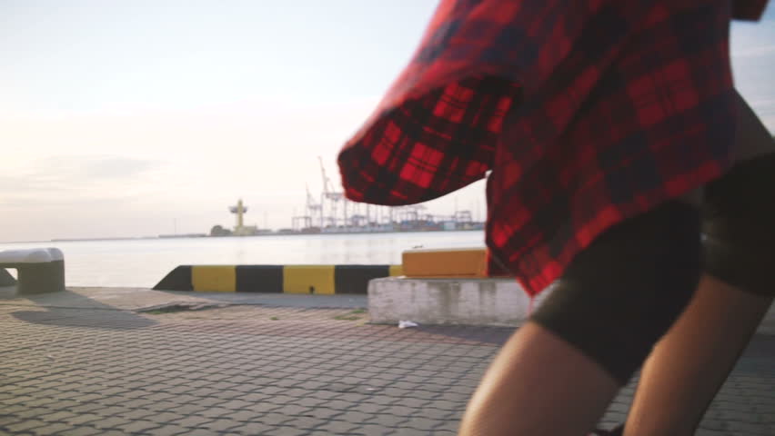 Young stylish funky girl with green hair riding roller skates and dancing near sea port during sunset, slow motion