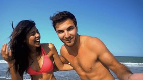Portrait of smiling multi ethnic couple in swimwear embracing and sending air kiss on the beach