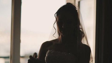 Young bride silhouette in white wedding dress and vail sprinkles parfume on hands and around room in front of window