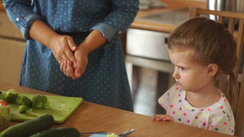 Little girl in the kitchen with her mother. Mom gives the daughter broccoli. The girl resolutely repels the vegetables with her hand. The baby is very angry and does not want to eat.