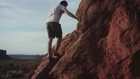 Bearded hipster man climbing red rock, rock formations in distance, Arizona or Utah desert, Summer 2017