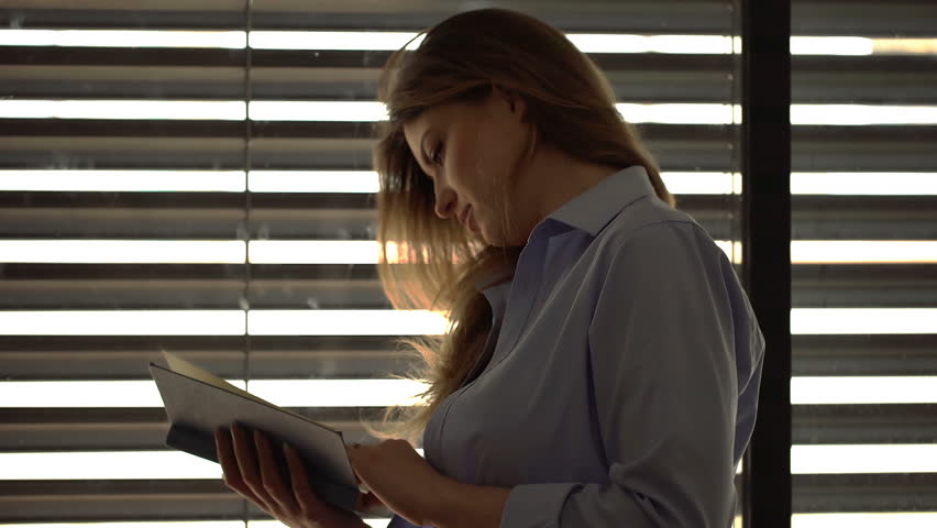 Attractive woman standing next to the window and reading book
