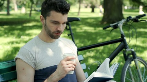 Handsome man drinking coffee in the park and reading book, steadycam shot
