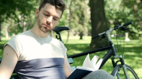 Handsome man receives message on smartphone while reading book in the park, steadycam shot
