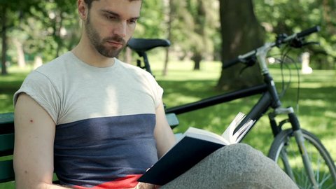Handsome man relaxing in the park and reading interesting book, steadycam shot
