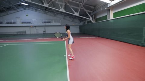 Full length portrait of slim sexy woman in sport outfit playing tennis at indoor court with professional equipment. Sportswoman serving and returning balls with racket jumping in recreation area.