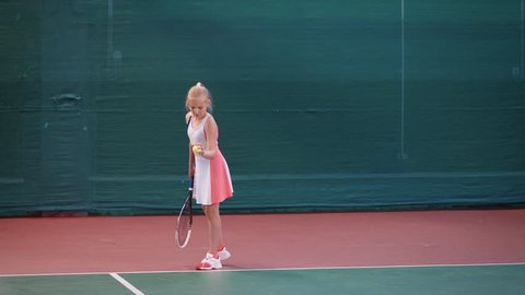 Little girl dressed in sport outfit is having training at indoor tennis court. Young female sportswoman is playing professional tennis and bouncing yellow ball before hitting it with racket.