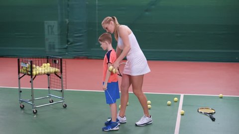Female coach dressed in short white sport dress is teaching little boy to play at indoor court. Professional tennis instructor is showing to male toddler process of returning a ball with racket.