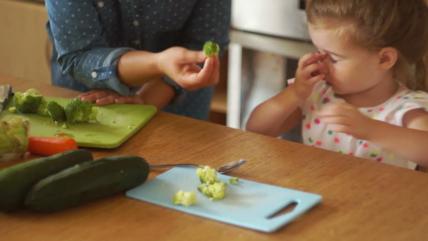 Portrait of a little girl in the kitchen. Mom gives the daughter a broccoli and a carrot. The girl pushes the vegetables away. Healthy food. Childish whims. Royalty-Free Stock Footage #29468866