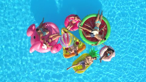 AERIAL TOP DOWN Cheerful girls and guys playing with ball on colourful floaties in pool. Smiling friends enjoying summer vacation on inflatable pineapple, pizza, flamingo, watermelon, doughnut floats