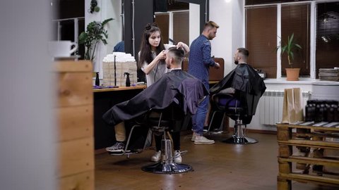 Image of interior of barbershop and two professional hairstylists. Female and male barbers working with clients sitting inside. Woman and man using instrument making perfect shape for haircuts.