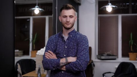 Portrait of young handsome man dressed in casual shirt posing in stylish modern interior of barbershop. Confident stylist standing near his workplace folding his arms on chest looking at the camera.