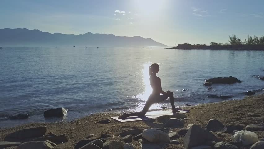 side view girl silhouette stands in yoga pose on rocky beach against pictorial rising sun reflection