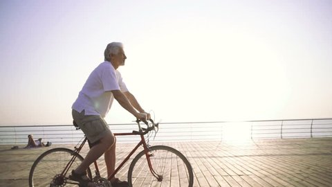 Handsome senior man riding bike on seafront, slow motion